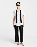 A model stands against a light gray studio background wearing a white short-sleeve button-up shirt with two wide black side panels, black wide-leg pants, black strappy sandals, and black sunglasses.
